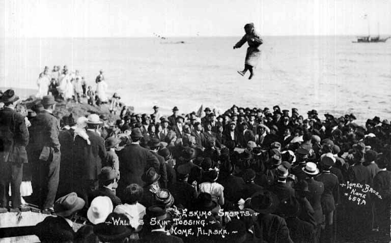 A vintage black and White photo of a large group of people surrounding a person who is jumping, or being tossed, on a walrus skin trampoline. We cannot see the trampoline but we see the jumper in traditional fur clothing, aloft high above the crowd. The crowd is assembled on a beach and there are two boats in the water.