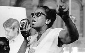 A black and white photo of Ella Baker, A dark skinned, feminine presenting person with black hair. She is wearing sunglass, and is speaking into a large microphone that she is holding. Here right fist is raised, as if to emphasize her message.