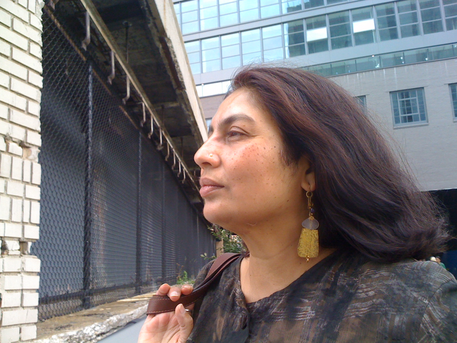 A headshot of Chandra Talpade Mohanty, brown-skinned feminine presenting person with red-brown hair. She is wearing large gold earrings and a small nose ring. She is in profile, and appears to be in motion with her the strap of a handbag tossed over her shoulder and her hair blowing in a breeze.