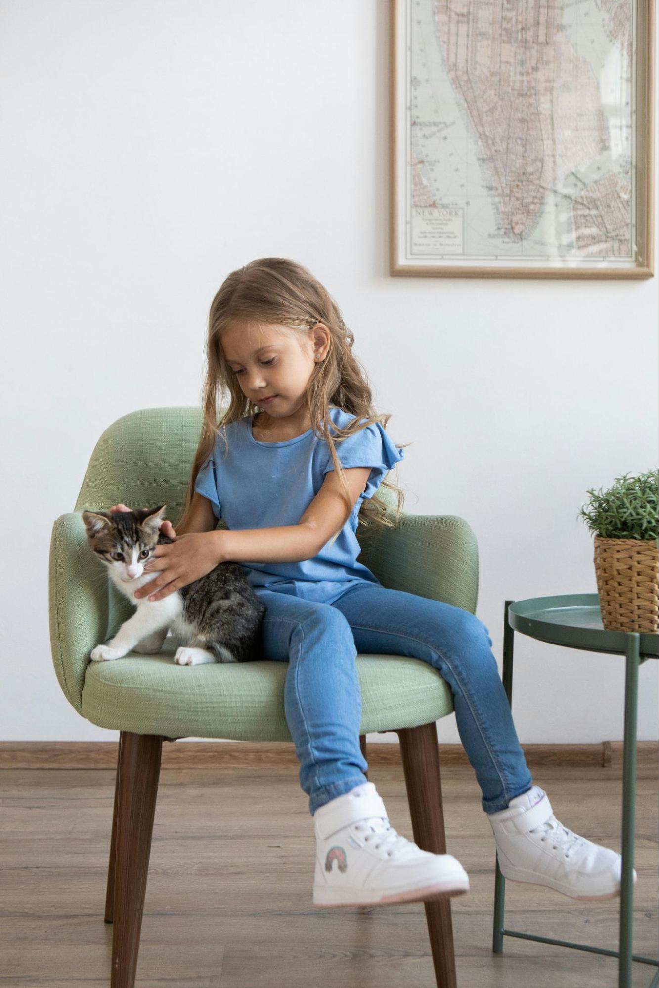 A feminine presenting child siting in a chair, playing with a small striped kitten.