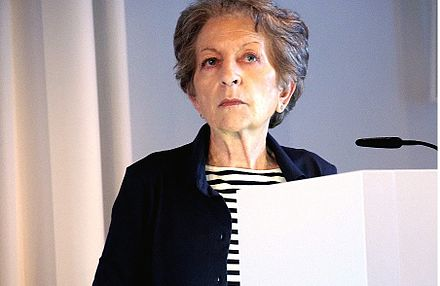 A head shot of Teresa de Lauretis, an older, light-skinned feminine presenting person standing at a lectern. She has short grey hair and is waring a blue and white striped shirt and a blue jacket.