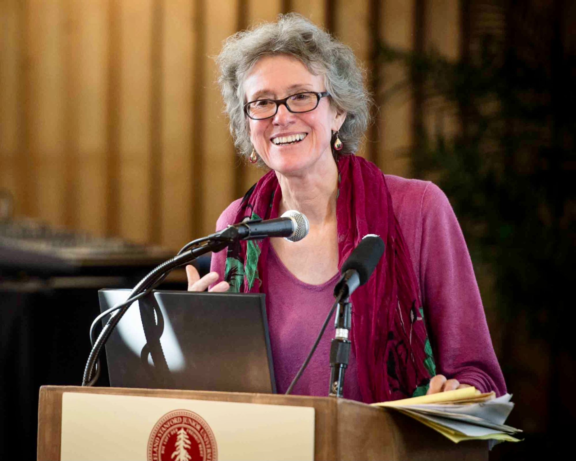 A headshot of ArlieArlie Russell Hochschild, an older, light-skinned, feminine-presenting person, who is standing at a lectern. she has grey hair and glasses, and is wearing a pink blouse and a red scarf. She is smiling.