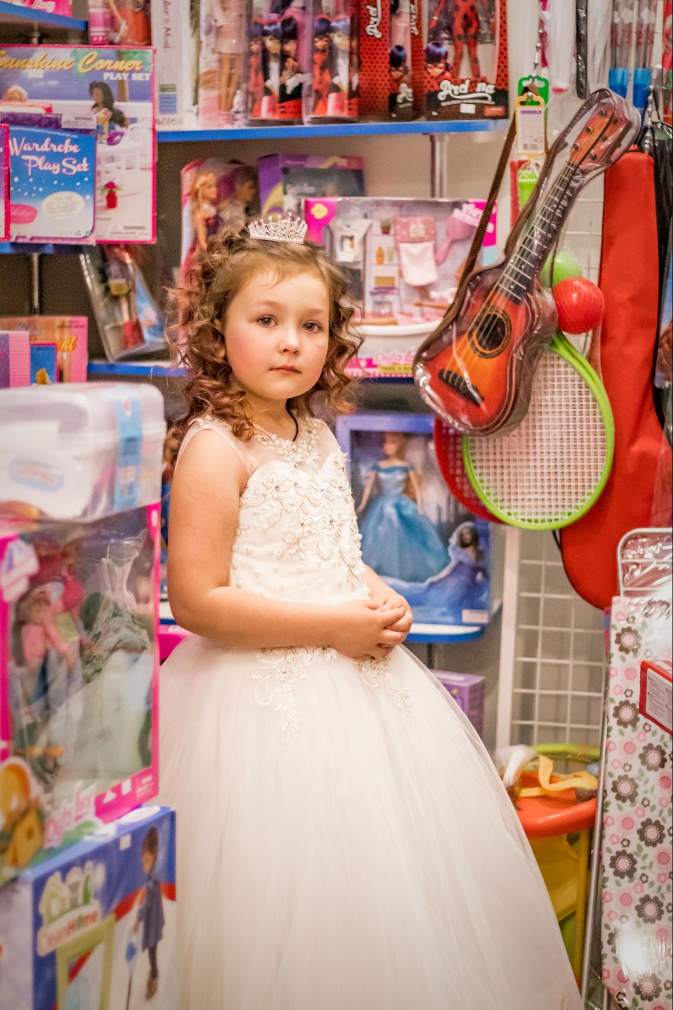 A young, blond, light-skinned femme presenting child in a the toy section of a store. She is wearing a white princess costume with a silver crown. The packaged toys on the shelves include dolls, makeup kits, and musical instruments. The only sports equipment is a wiffle ball set.