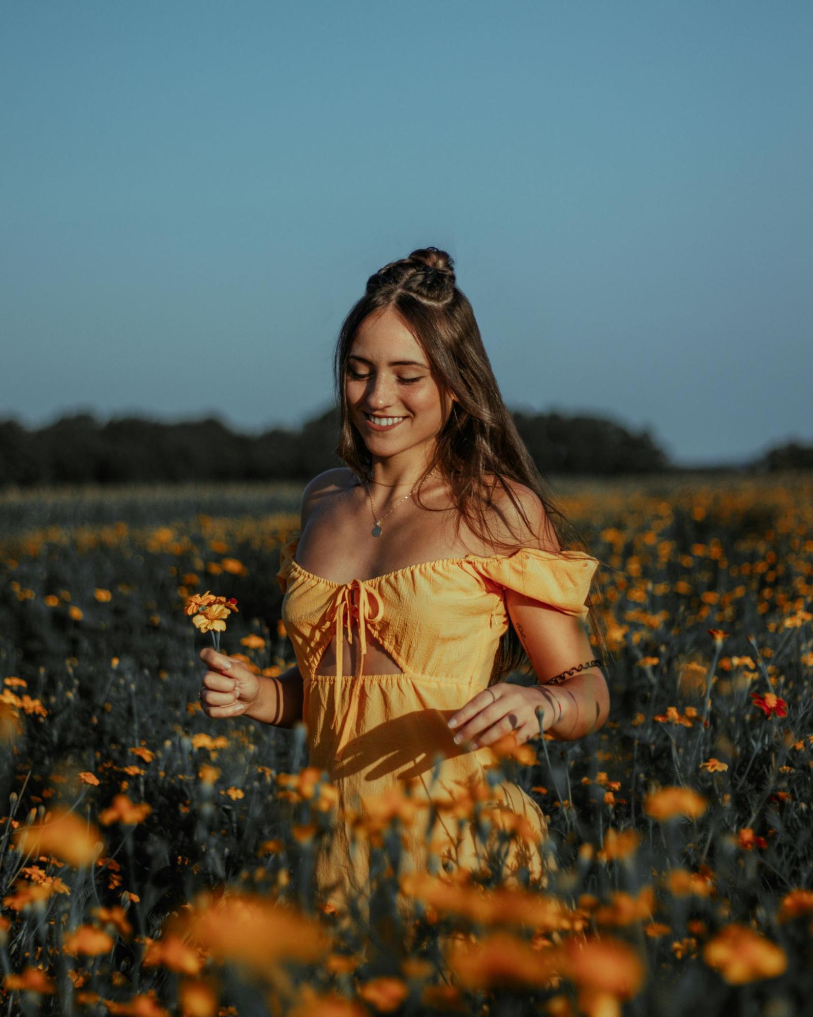 A young, thin, light-skinned woman with long hair and an off-the-shoulder dress smiles in a field of flowers