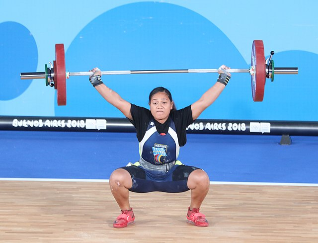 The power lifter, Maximina Uepa (she/her) doing a deadlift. she is feminine presenting person with light brown skin, and textured black hair in a low ponytail. She is wearing black spandex shorts and a t-shirt under a blue and white vest that says, "Nauru."