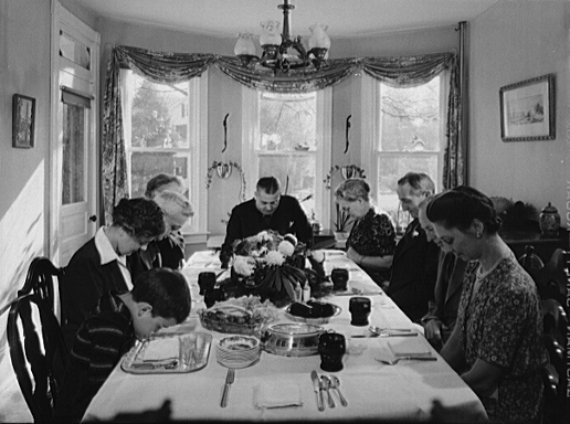 A family of light-skinned people bow their heads in prayer around a long table laden with food in a well-furnished dining room