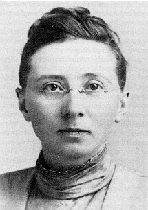 Black and white headshot of a young woman with dark hair. Her hair is in a simple braided bun on top of her head. Her clothing is from the Edwardian era and she is wearing glasses.