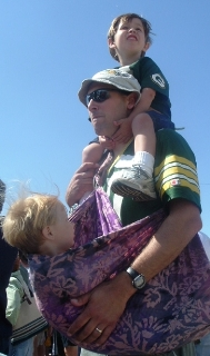 A light-skinned man with two light-skinned young children. One child is on his shoulders, and the other is in a purple batik baby sling. The dad and the child on his shoulder are both wearing Green Bay Packers shirts.