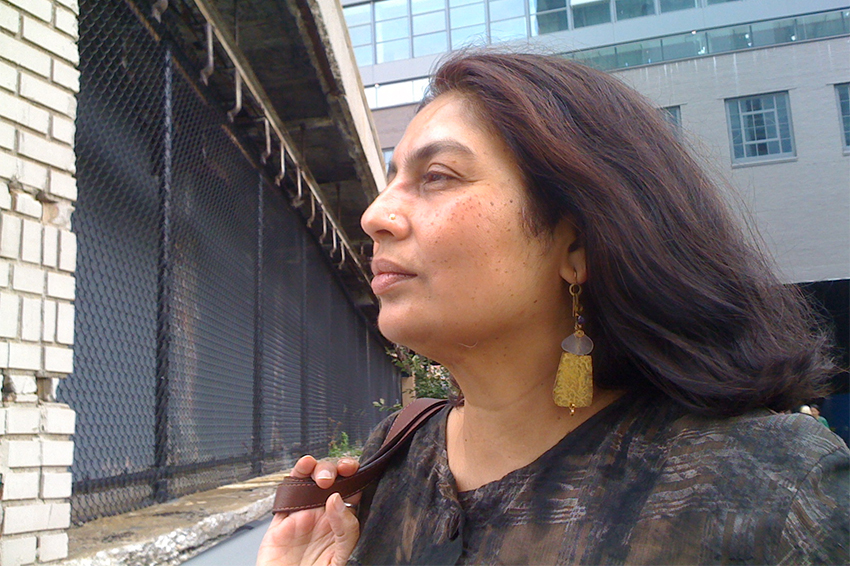 Chandra Talpade Mohanty A headshot of Chandra Talpade Mohanty, brown-skinned feminine presenting person with red-brown hair. She is wearing large gold earrings and a small nose ring. She is in profile, and appears to be in motion with her the strap of a handbag tossed over her shoulder and her hair blowing in a breeze.