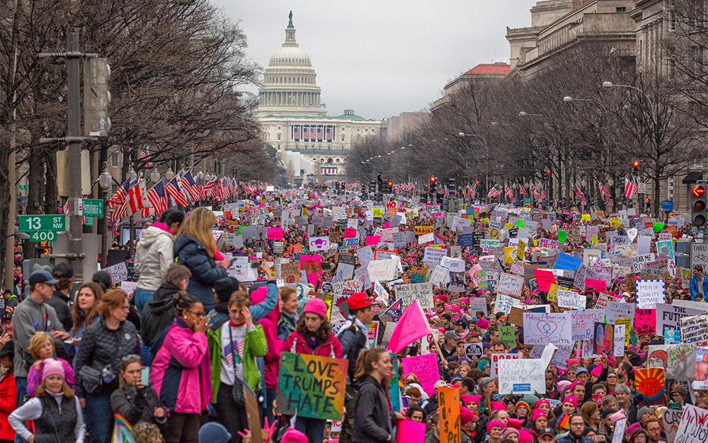 The 2017 Women’s March A very large group of people carrying signs and banners at the 2017 Women's March. Many are wearing pink hats. The Capitol Building is in the background.