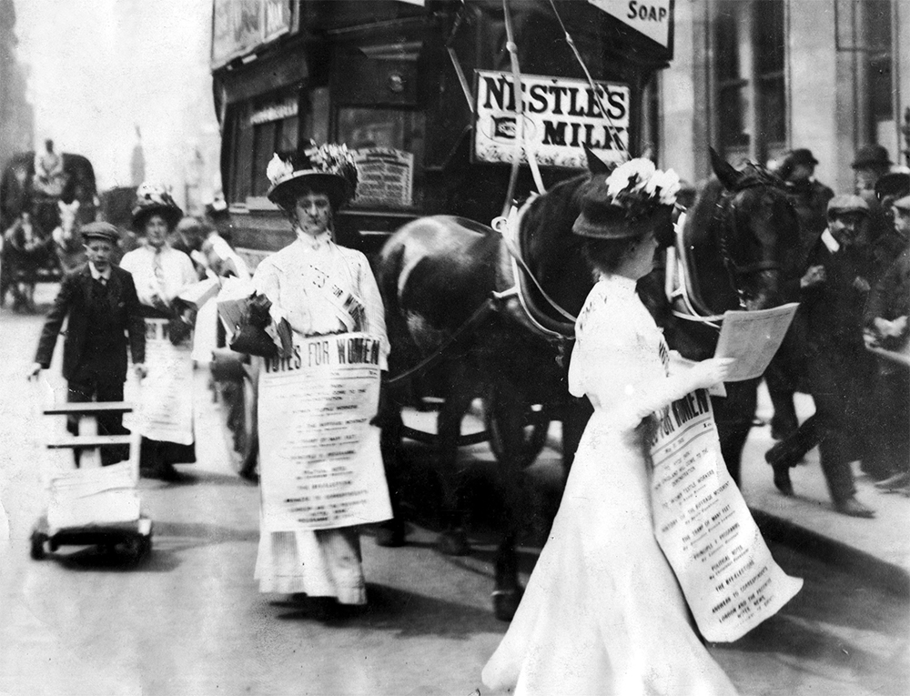 Votes for Women Grayscale photo from the 1900s of women in white dresses passing out fliers. They are wearing banners that say 'Votes For Women.'