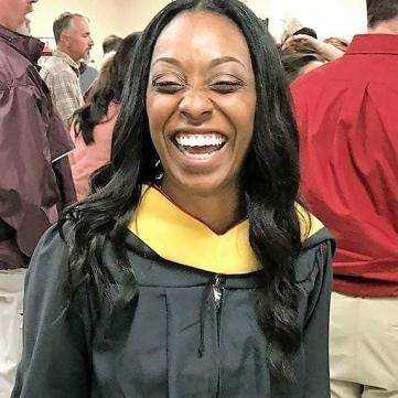 Aikia, a person with medium dark skin and long flowing hair, smiles in graduation regalia