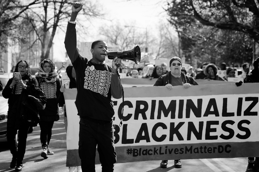 A young back man holding a bull horn wearing a shirt that says Black Lives Matter at a protest outside. People behind him carrying signs.