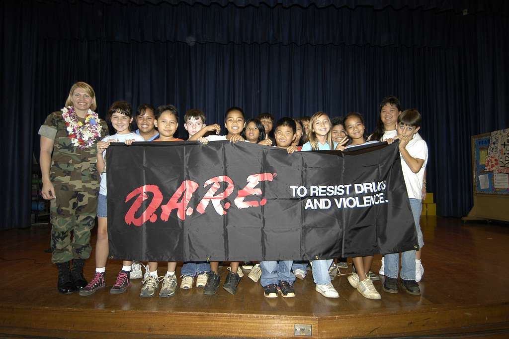 A woman wearing camouflage stands next to a group of kids holding a DARE banner