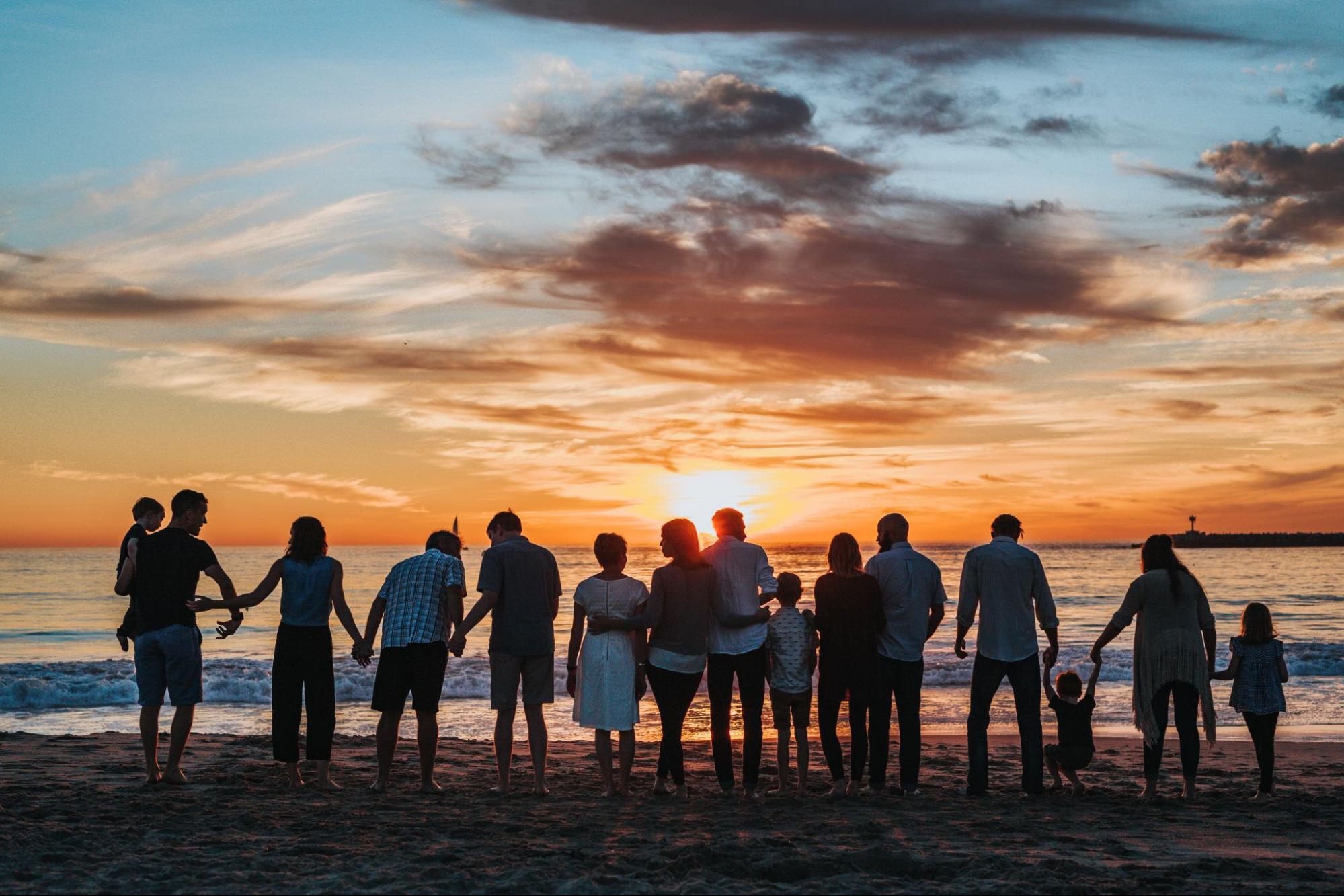 A large group of people silhouetted in front of a sunset on a beach, holding hands and hugging.