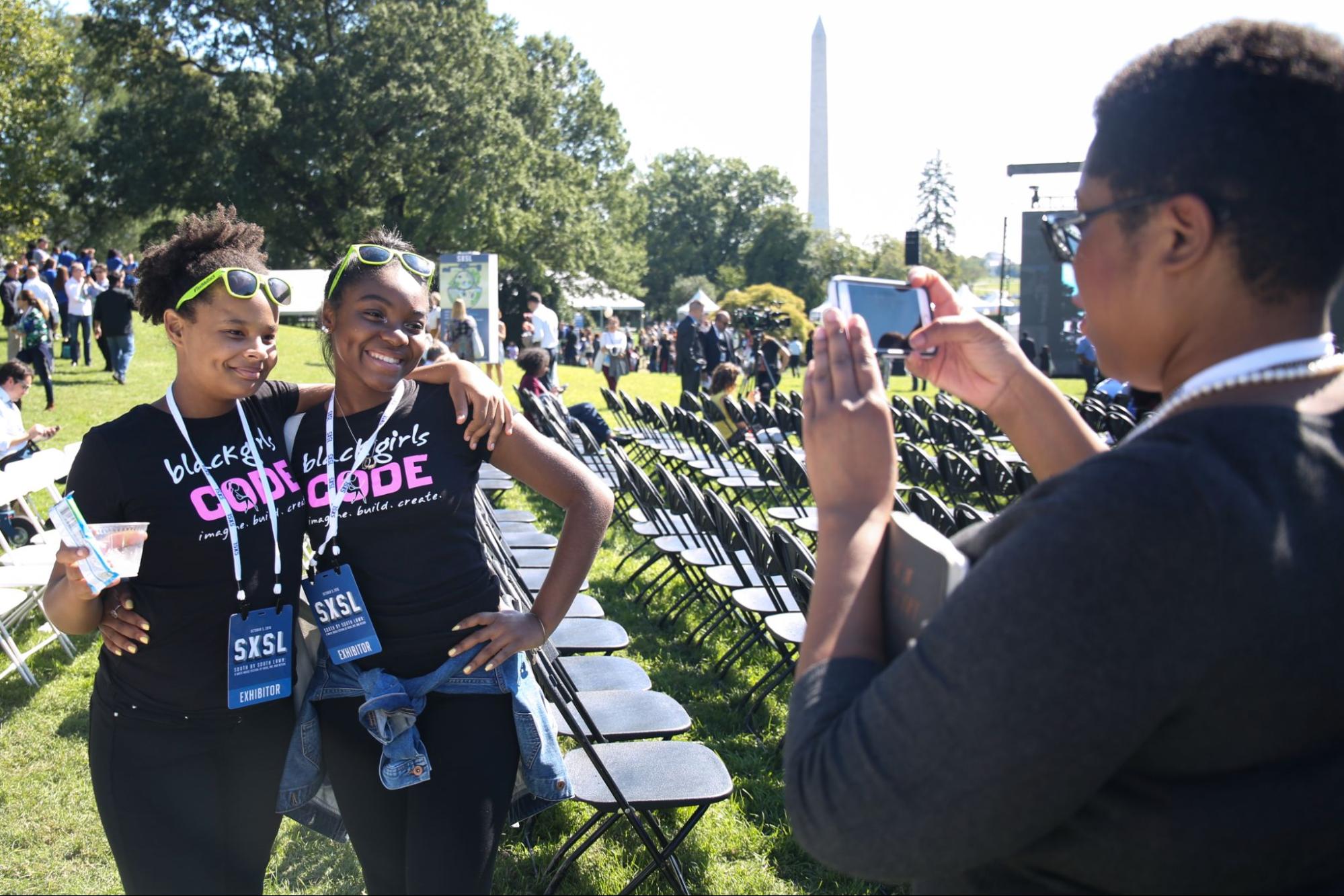 Two young dark-skinned women posefor a cellphone photo at an outdoor event wearing Black Girls Code tshirts.