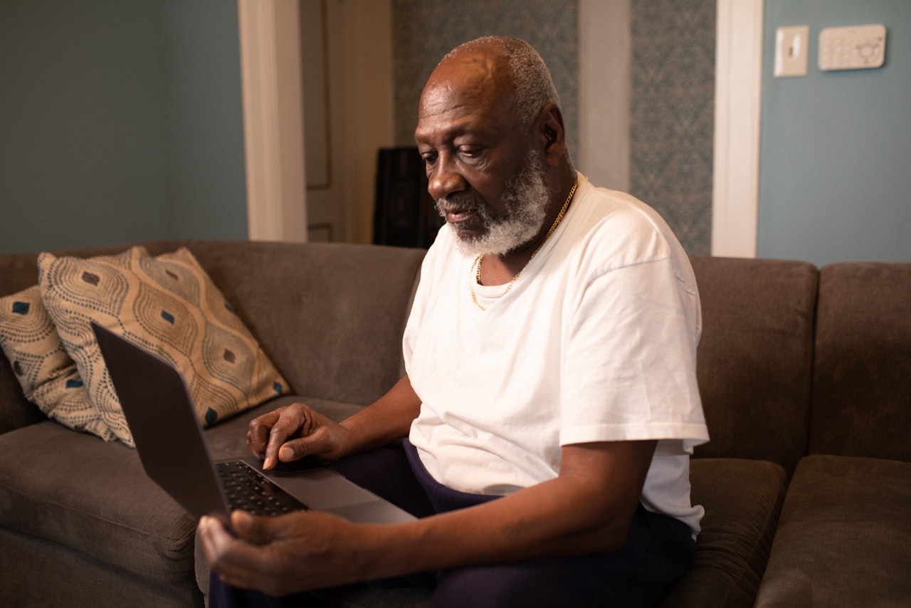 A Black man with a white beard sits on a couch with a laptop.