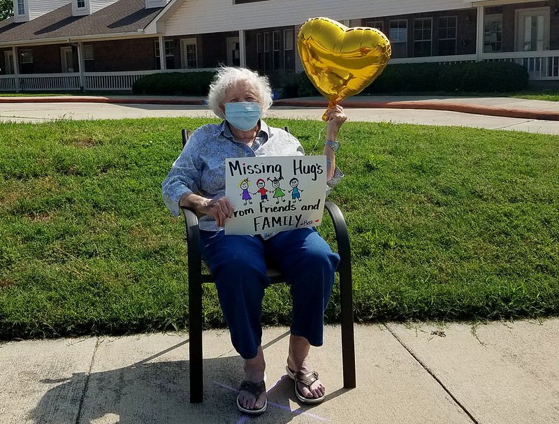 An older woman sits outside with a balloon and a sign that says "Missing hugs from friends and family"