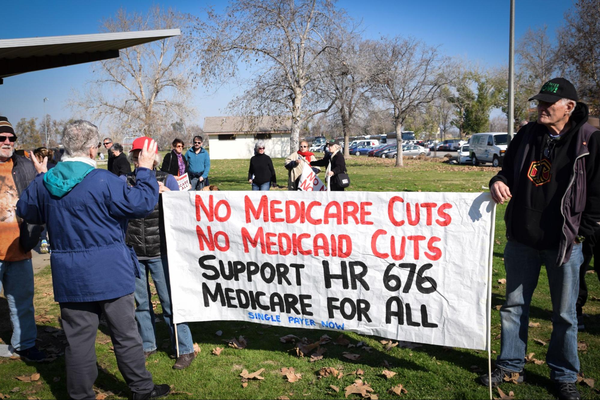Outdoor protest. Older people have a sign that says "No medicare cuts, no medicaid cuts, support HR 676, medicare for all, single payer now"