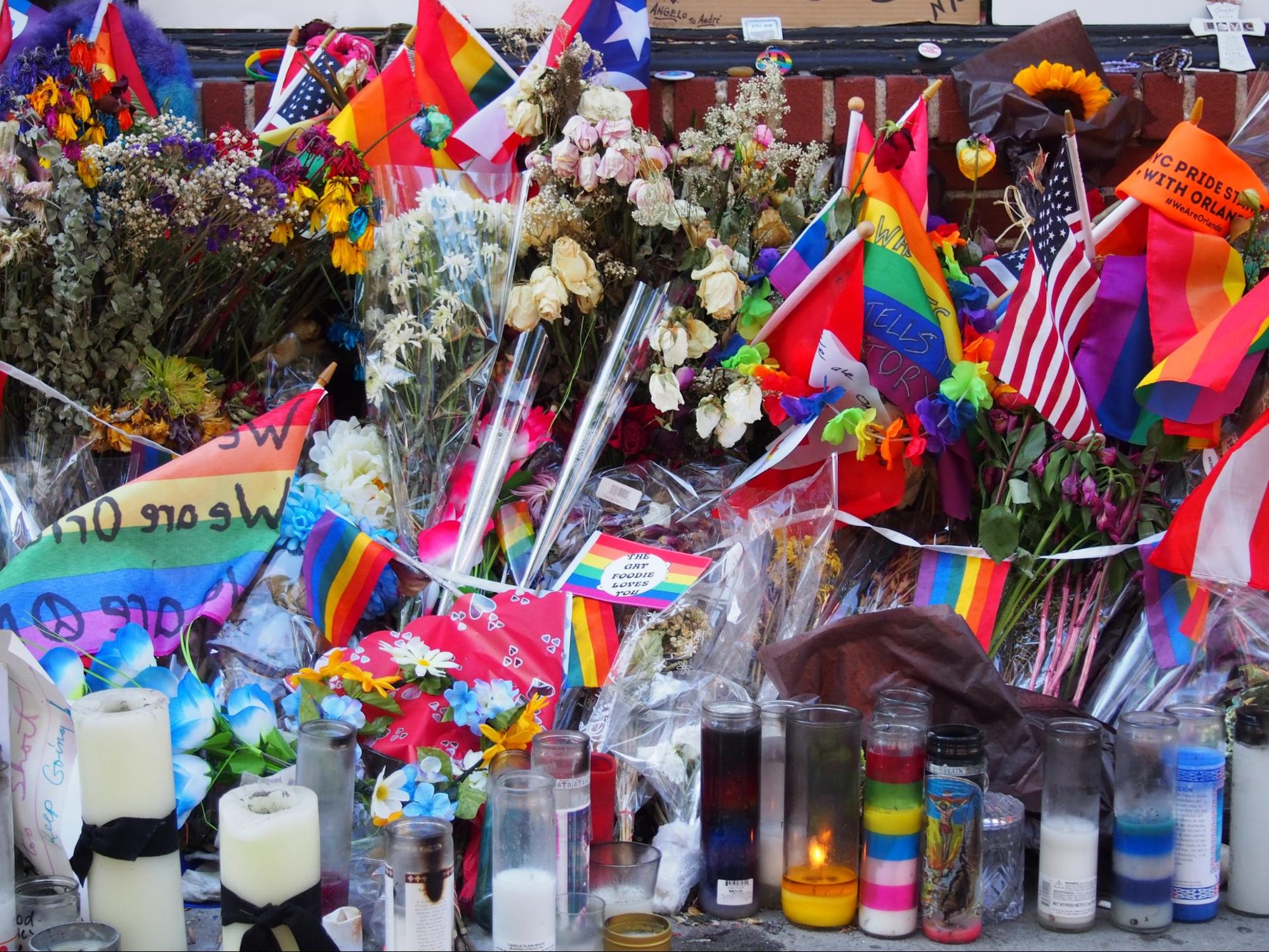 A memorial scene in front of the Stonewall Inn: rainbow flags surrounded by candles and flowers.
