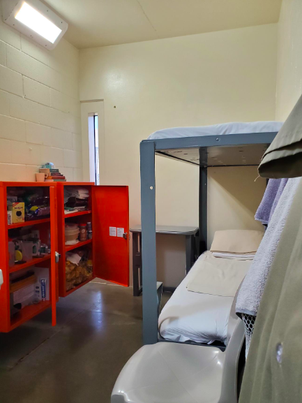 Inside of a prison cell with a bunk bed, a chair, and 2 red cabinets of personal items.