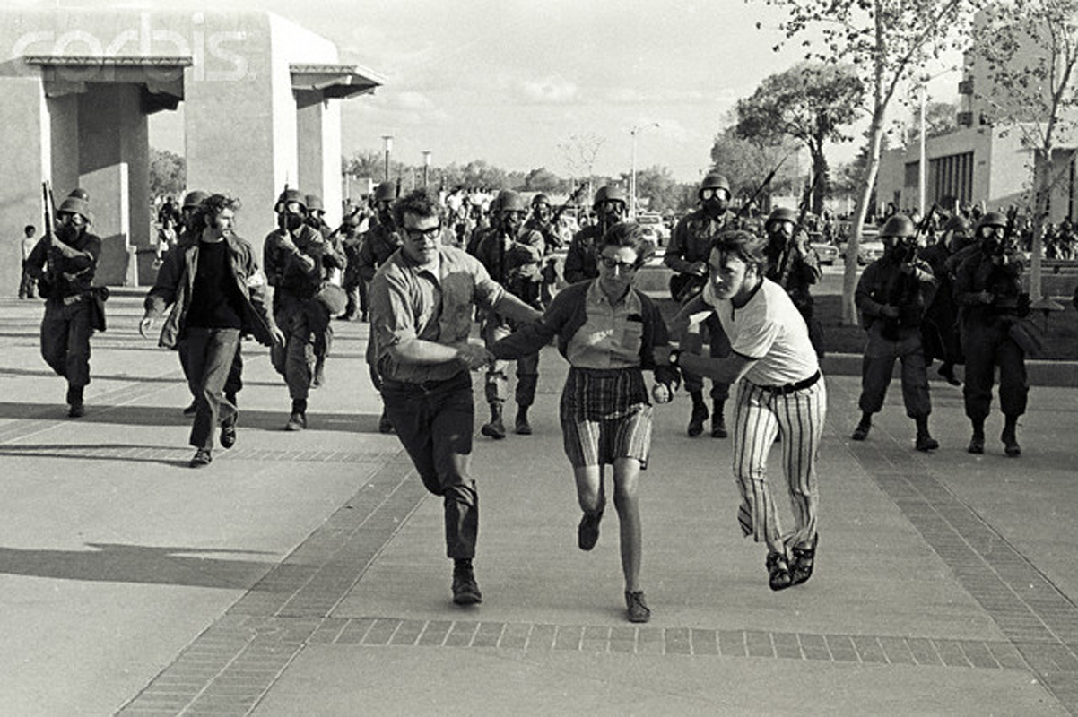 a black and white photograph of people with light skin running from a line of police. Two of them are men, helping a woman run.