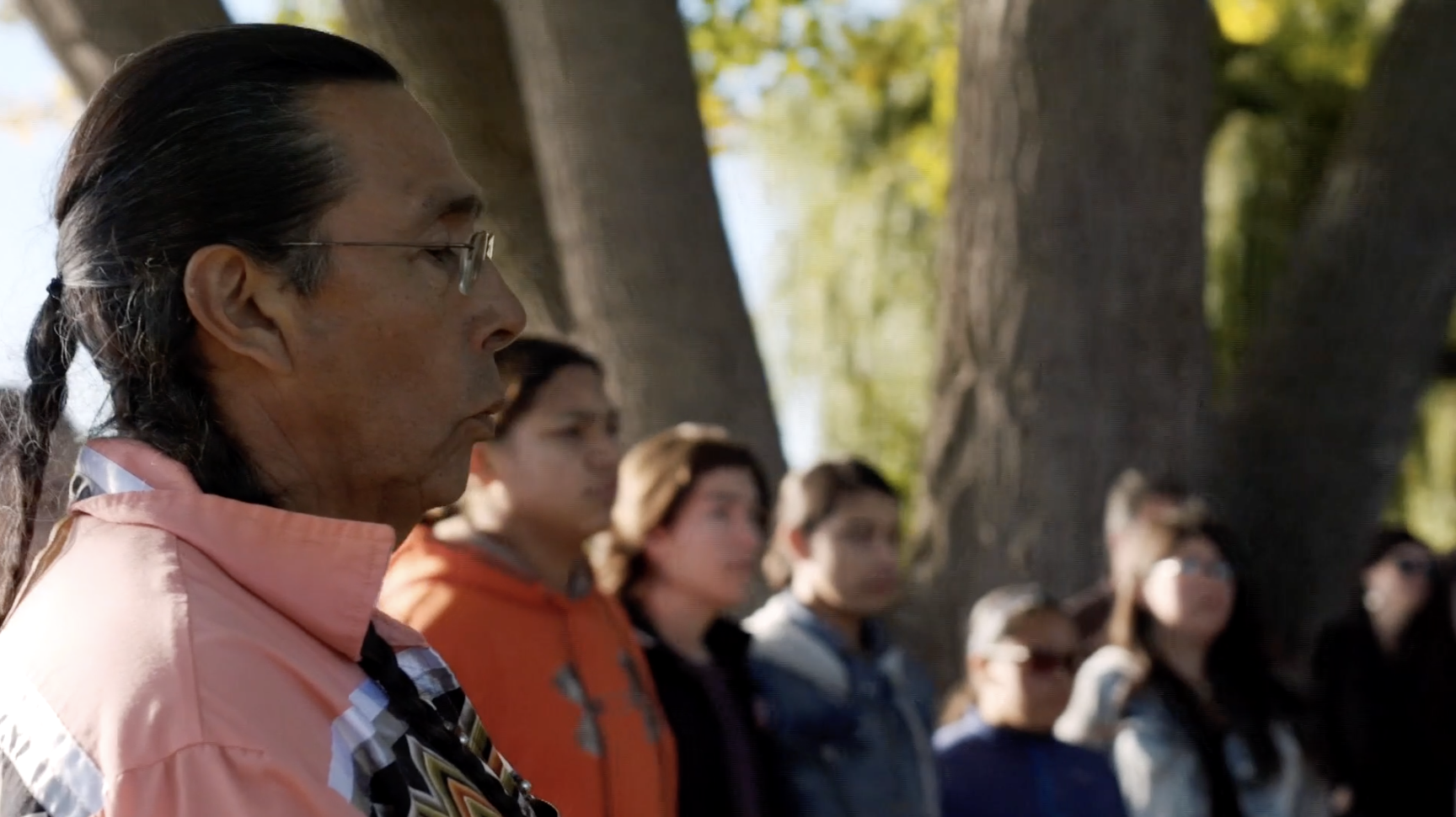 screenshot of a man with brown skin, glasses, and long hair listening alongside others who are standing in a circle.