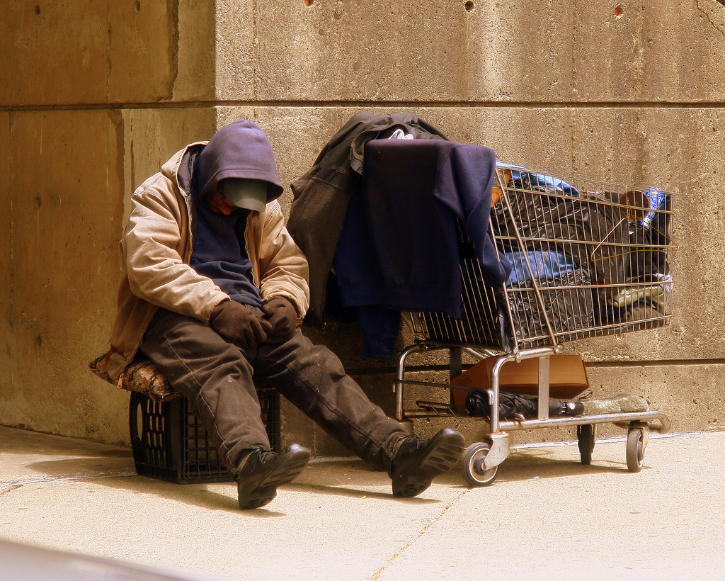 image of a person in very worn clothing, slumped on a plastic carton seat. A shopping cart filled with personal items sits nearby.