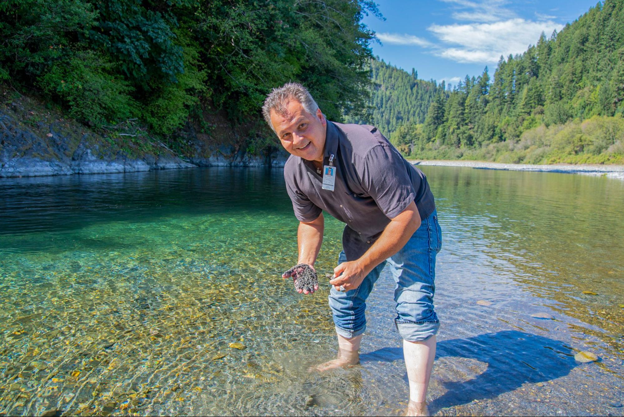 Smiling man wearing a purple shirt and rolled up jeans stands barefoot in the middle of a river. He is holding what looks like mud or silt from the river.
