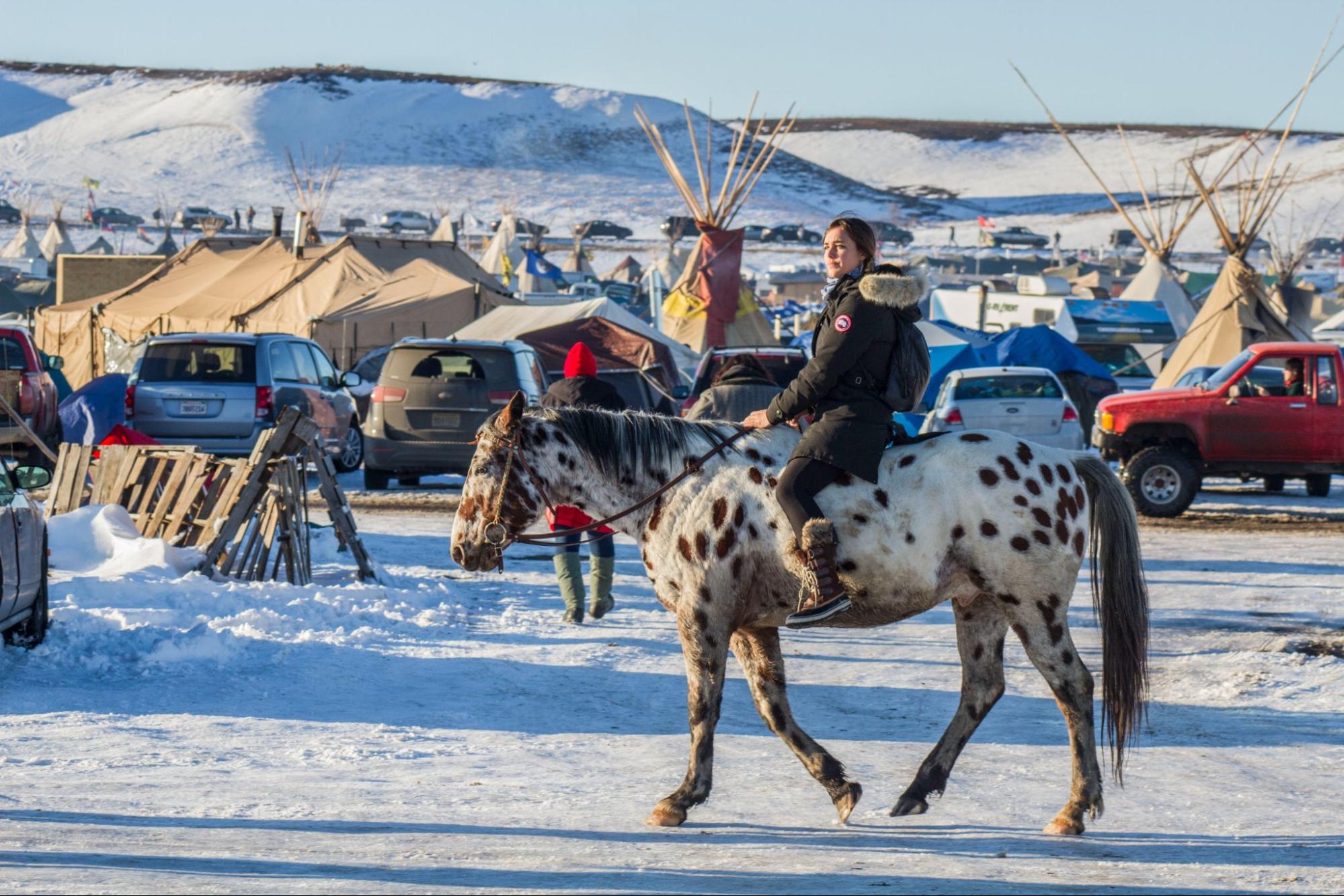 a woman rides a spotted horse in front of cars, tents and tipi. there is snow on the ground and in the hills nearby.