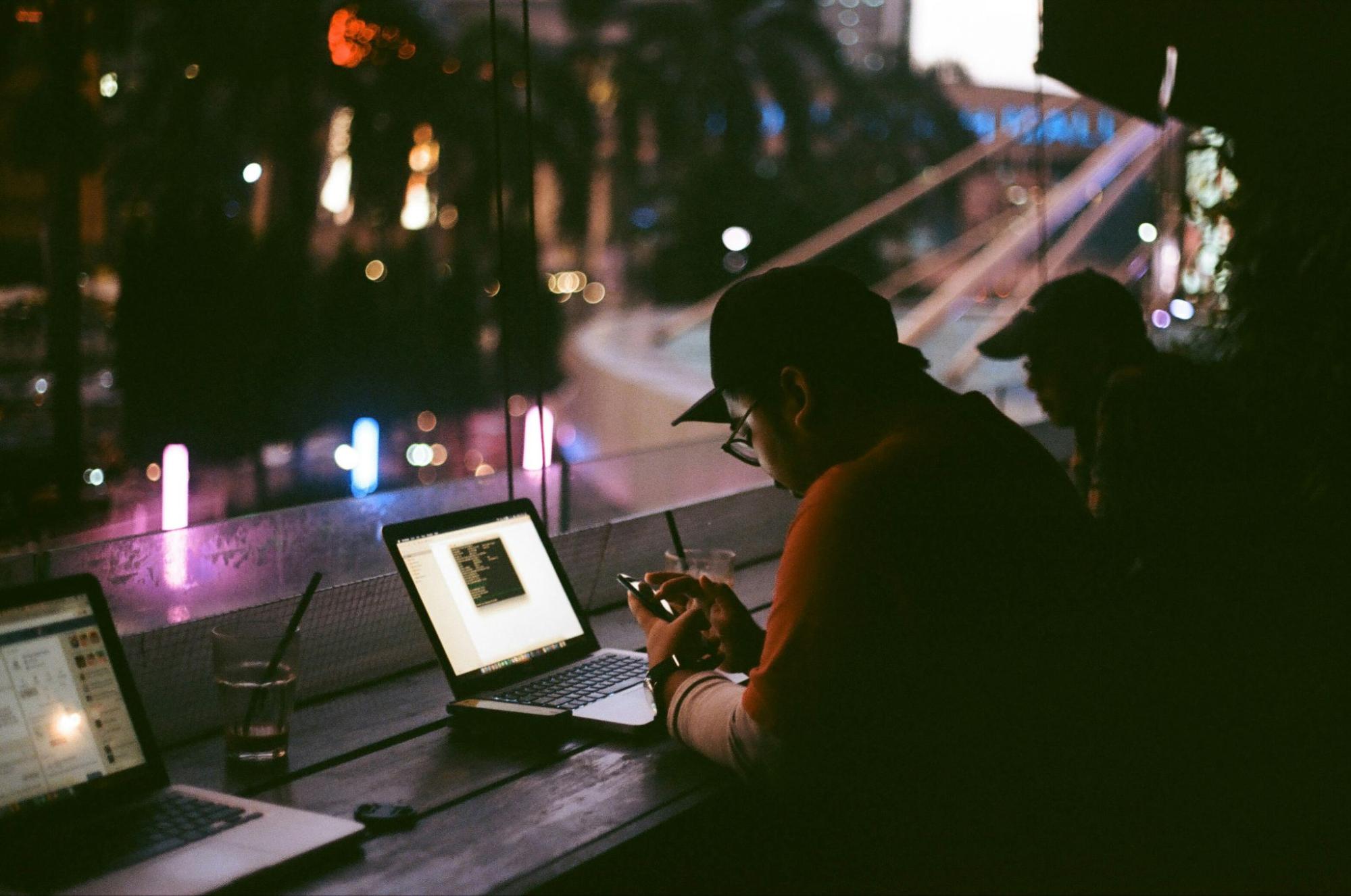photo of a man sitting in facing a window with night lights, in front of a computer and looking at his phone,