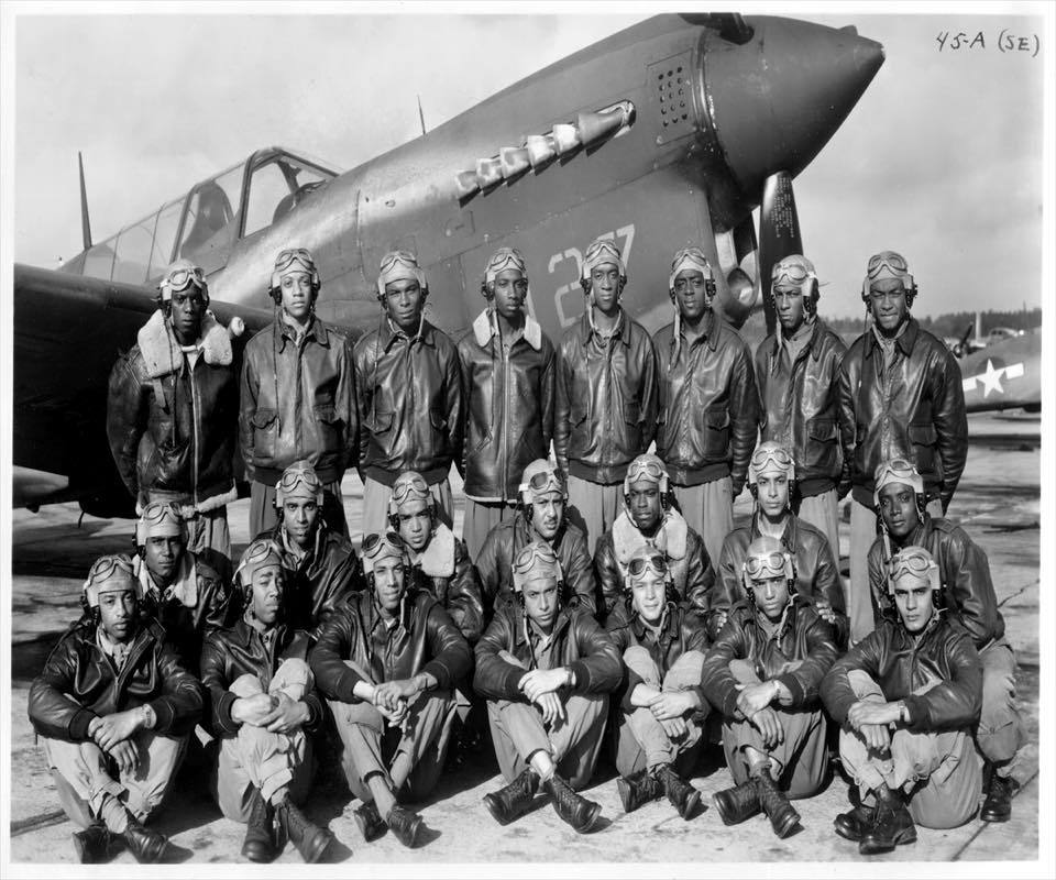 black and white photo of 22 dark skinned men in leather pilots' jackets standing in front of a old aircraft.