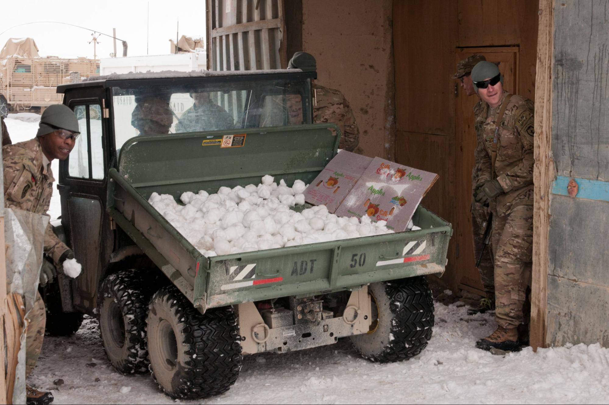 Two people dressed in camouflage stand poised, one with snowball in hand, beside fresh ammunition of snowballs collected in a truck.
