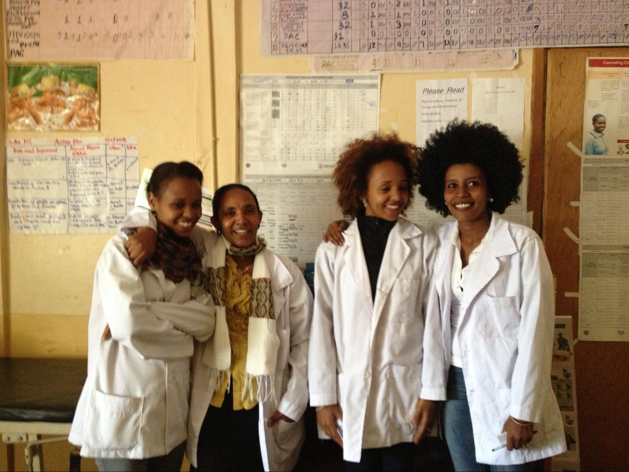 four dark skinned women in medical professional gowns stand in front of a classroom-like wall.