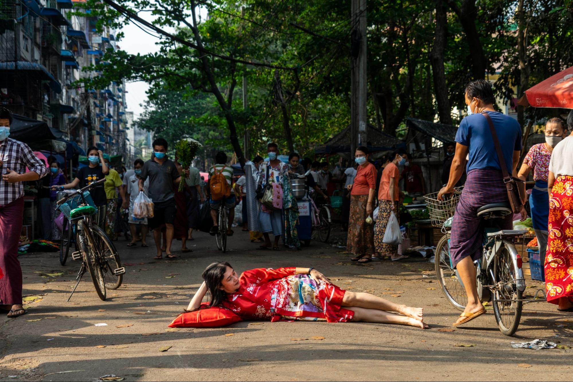 a woman dressed in a colorful red dress lies on her side in the street, her elbow rested on a pillow. Bystanders, some wiht bicycles stand by watching.