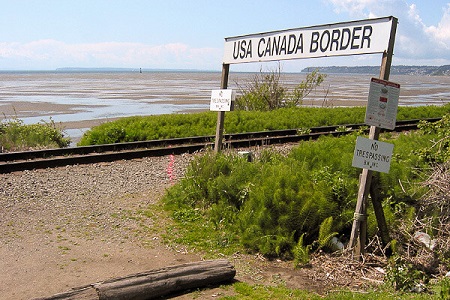photo of a sign that reads, USA Canada Border beside train tracks and a vast lanscape.
