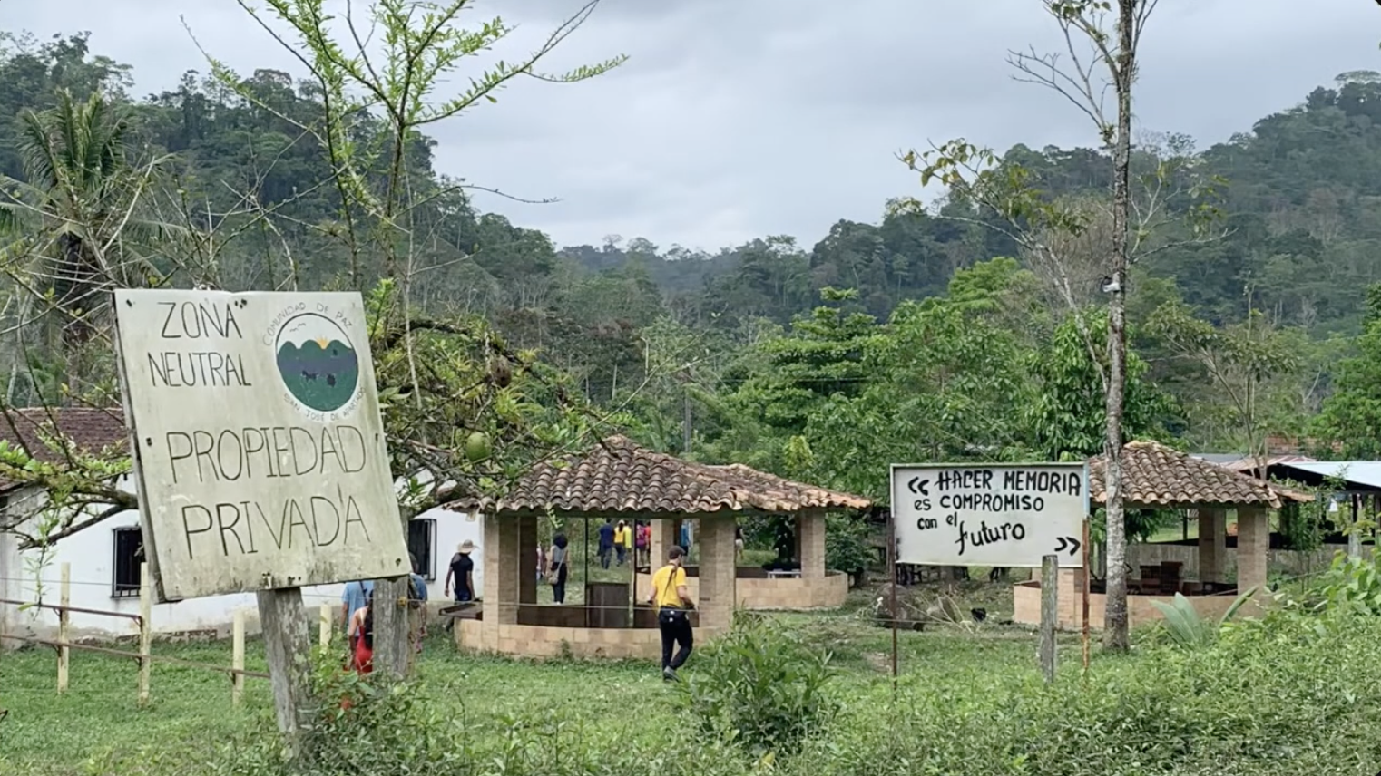 A series of small huts sit in a lush jungle with a few people walking under and around two signs.