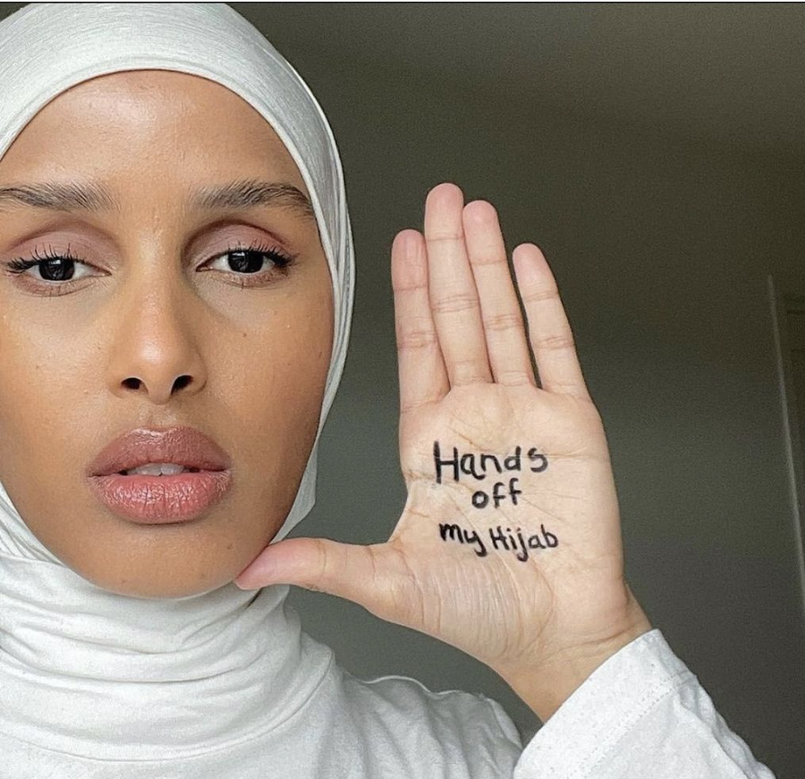 A Muslim woman wears a hijab that covers her head, neck, and arms. She holds her palm towards the camera with "Hands off my Hijab" written with black marker on it.