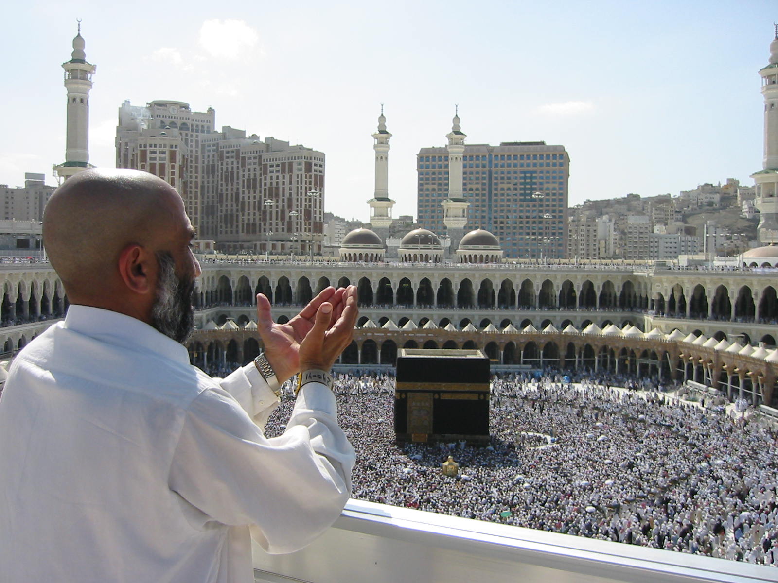 a dark skinned man in a white shirt stands above a very large crowd and Islamic architecture, with his hands, outstretched in prayer.