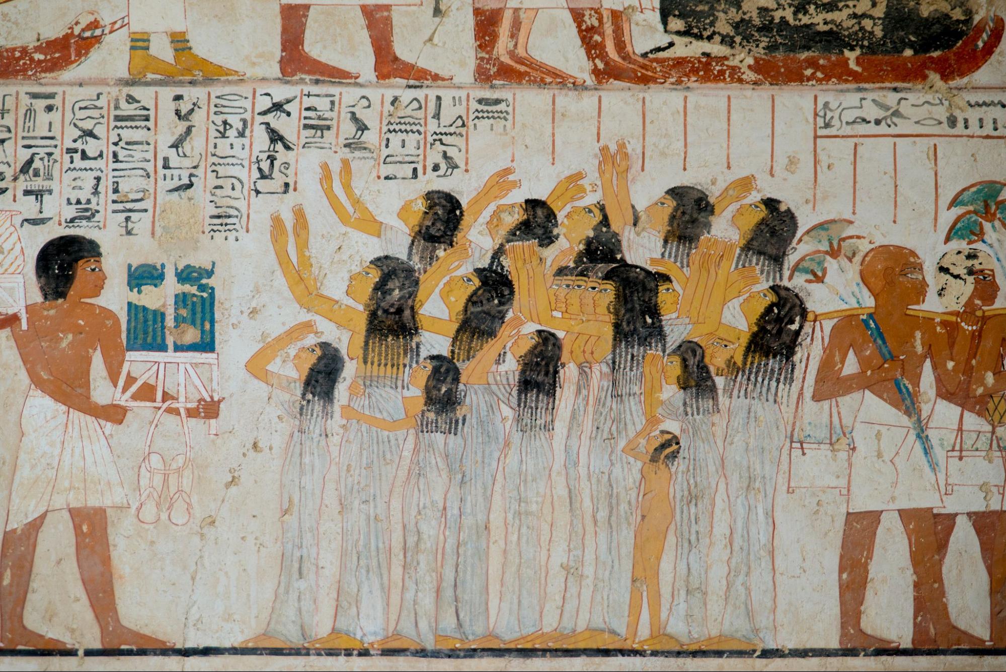 A photograph from an Egyptian tomb wall that shows women reaching towards the feet of people above them, men beside them holding fans, and carrying items with yolks.
