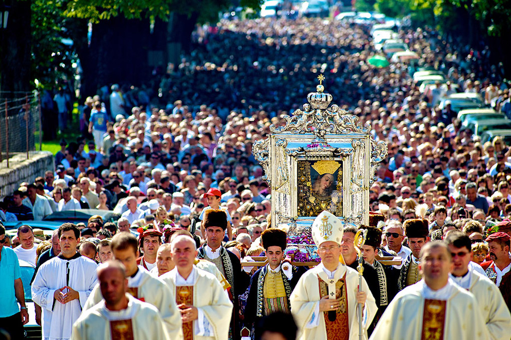 a photograph of a large crowd, walking down a street. men dressed in catholic garb are in front, a painting of the Virgin Mother Mary is carried behind them.