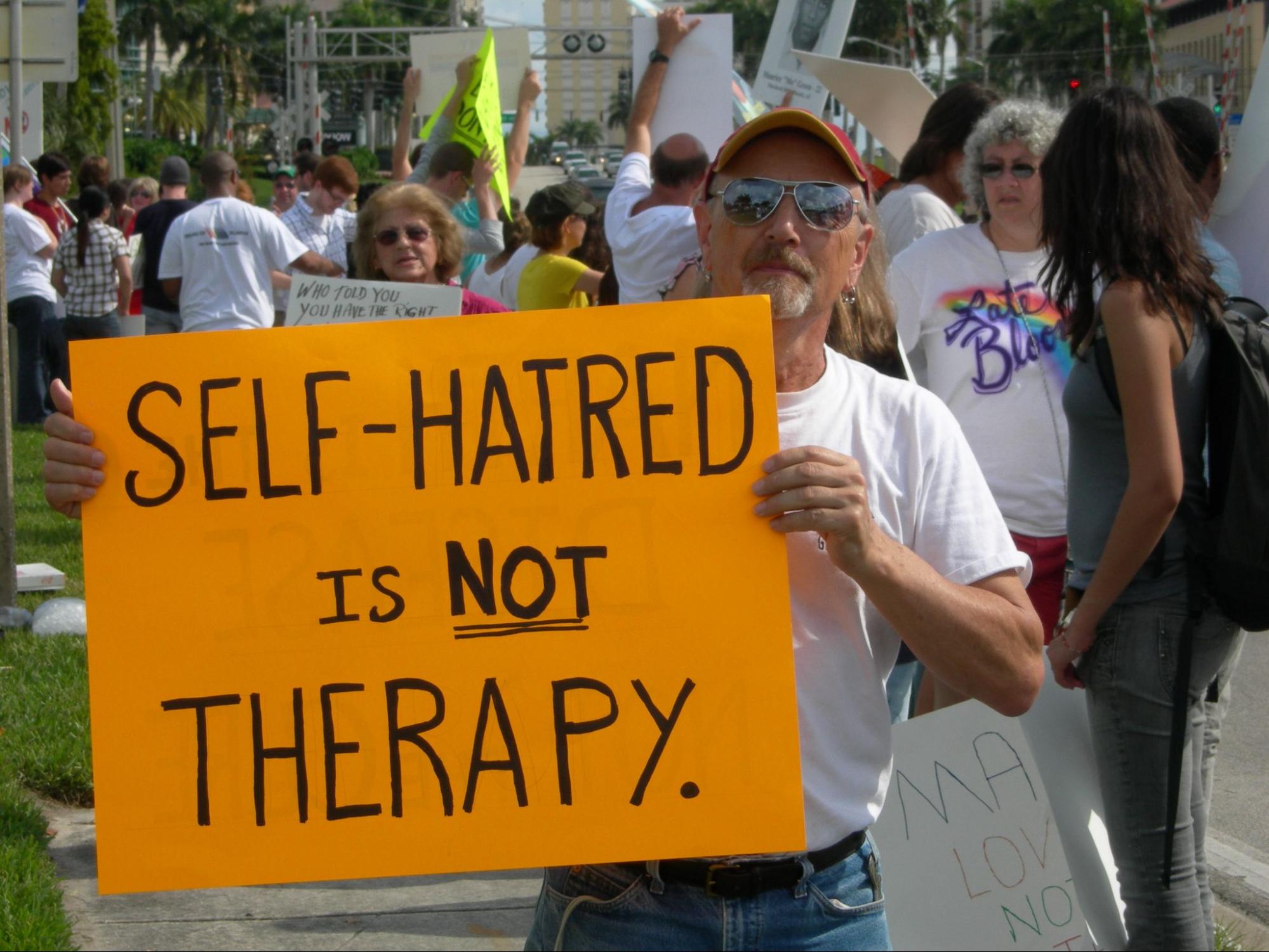 A large group of people participating in a protest. The focus is on a white man holding a sign that states "self-hatred is not therapy".