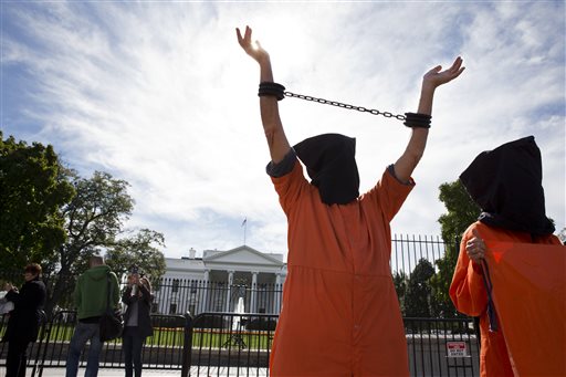 In front of the White House, a protester in a hood and jumpsuit lifts their arms high to show their shackles.