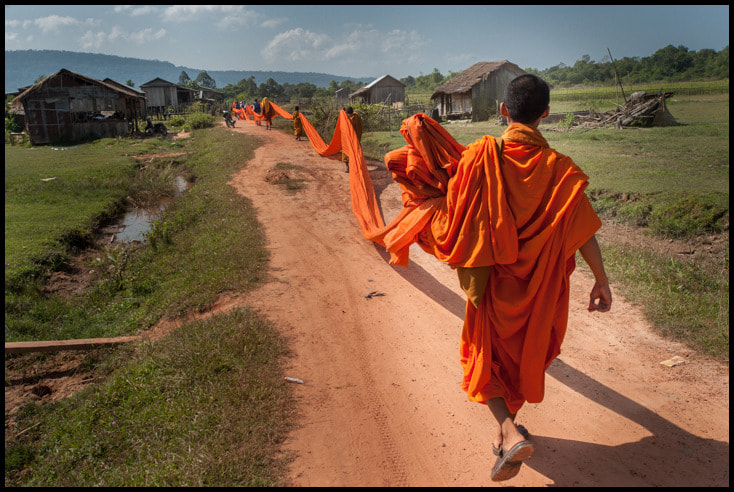 Buddhist monks in colourful orange robes march down a path holding orange fabric.
