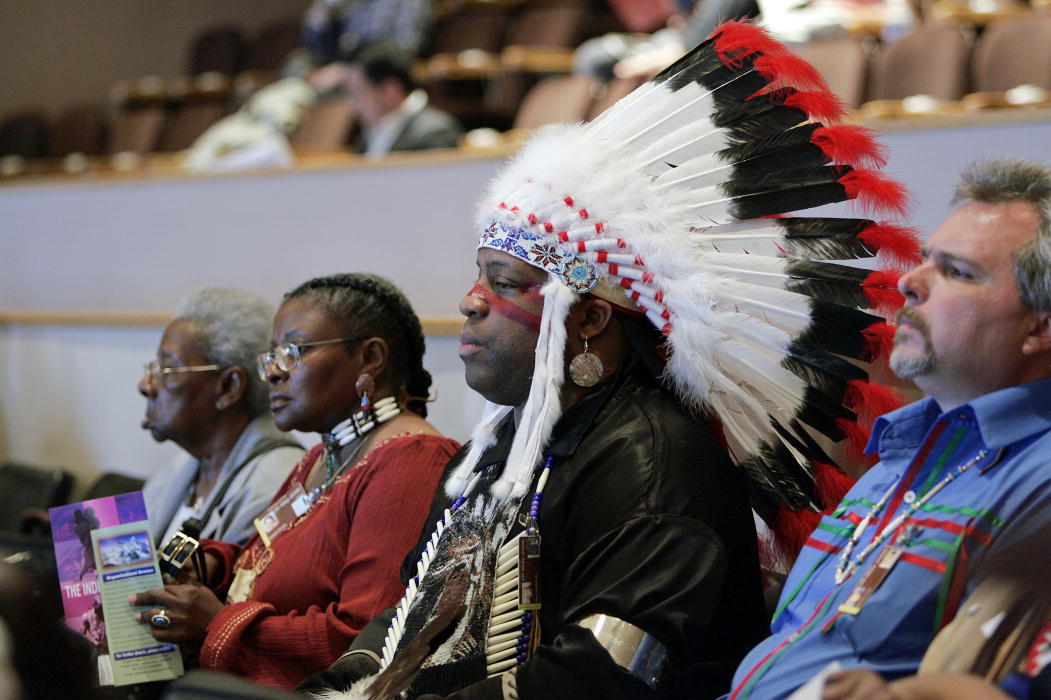 four people sit listening in a formal setting. one is a person with light skin wearing a colorful shirt. beside him is man with dark skin wearing a large headdress with feathers. beside him are two women with dark skin, one wearing native american jewelry.
