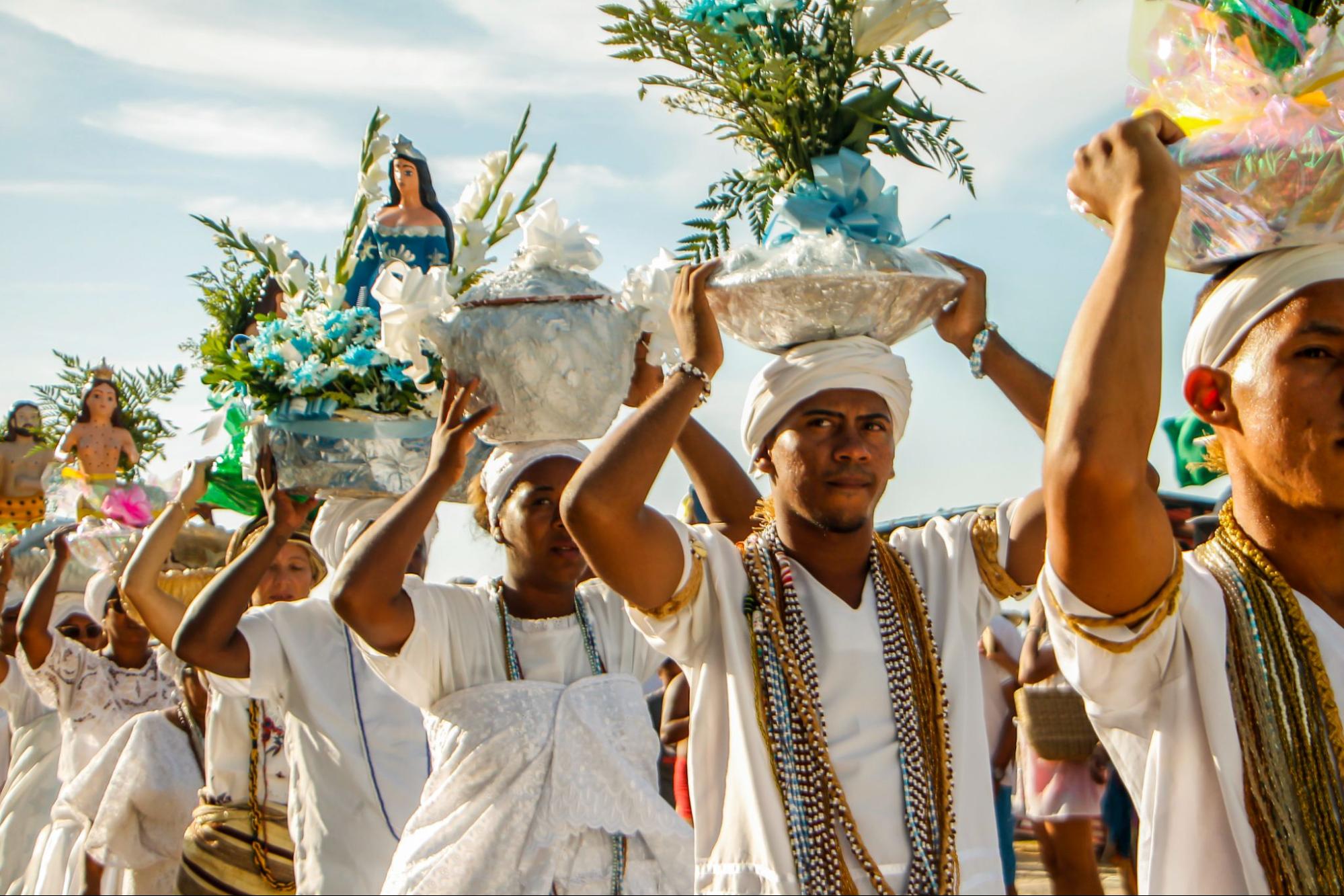 People in white cloaks and headdress, and neutral colored scarves walking with decorative bowls and statues on their heads.