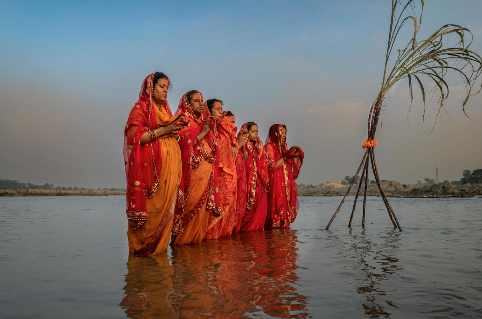 Six brown women in beautiful orange and red saris stand in the middle of the Ganges River. They look like they are praying.