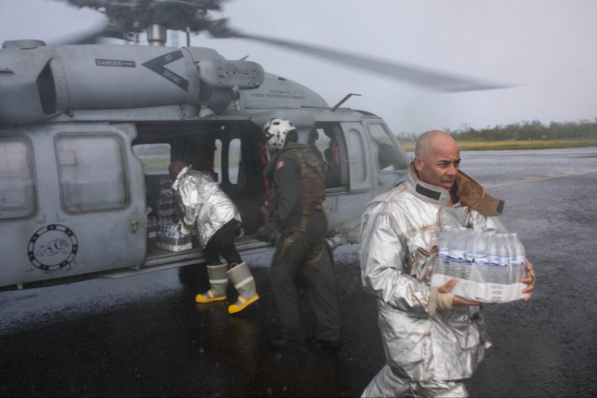 people in protective flight gear carry bottles of water away from a military helicopter.