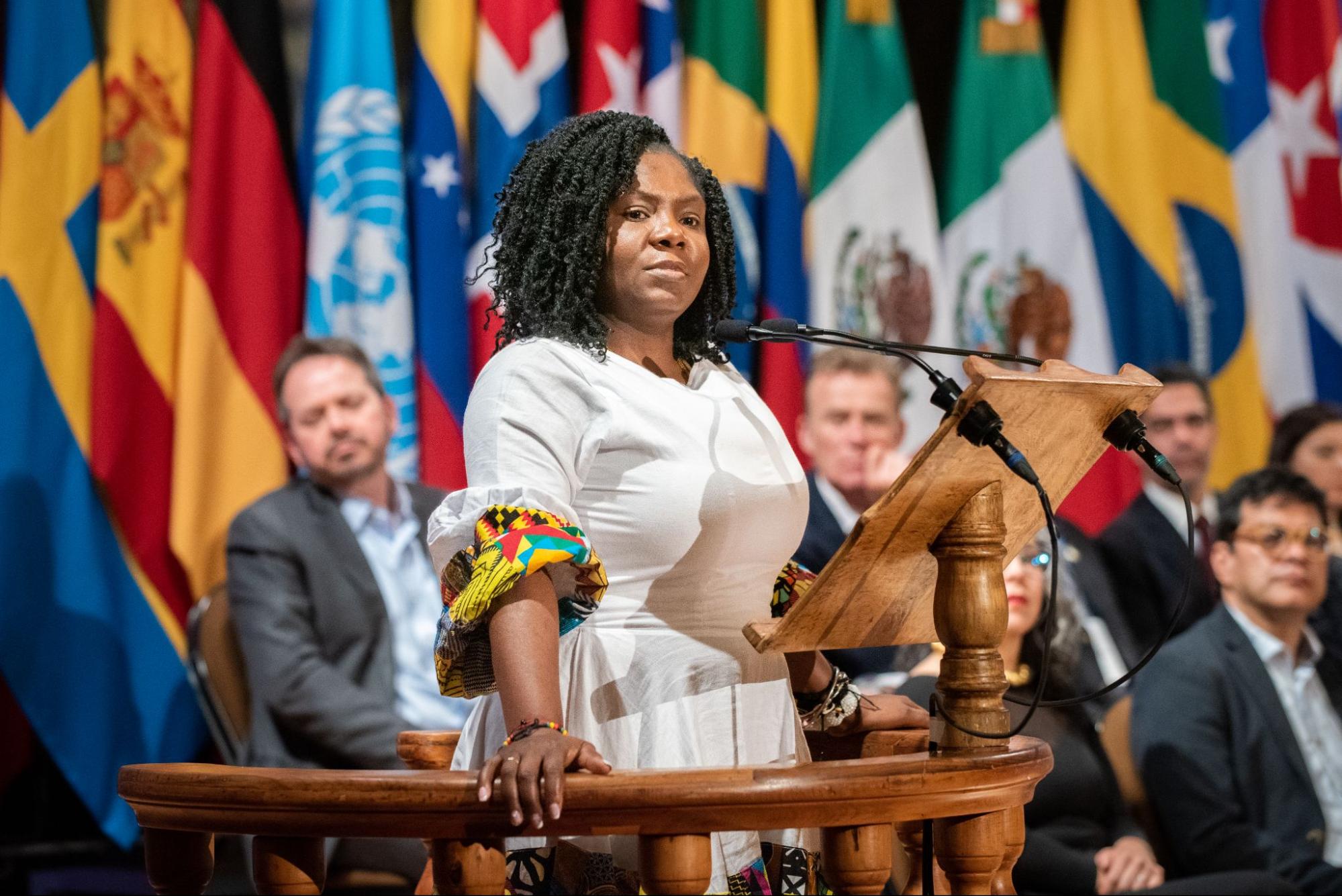 A dark skinned woman in a white dress with colorful sleeves stands in front of a microphone at a podium. Men in business suits sit behind her and in front of a wall of flags, listening.