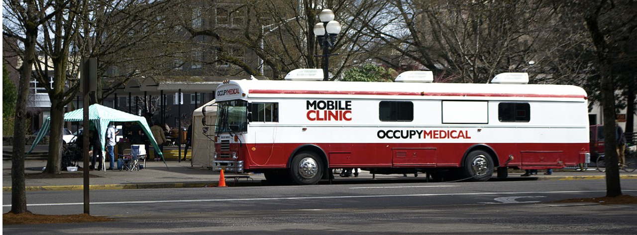 photograph of an extra-long bus parked on the street beside a distribution tent.